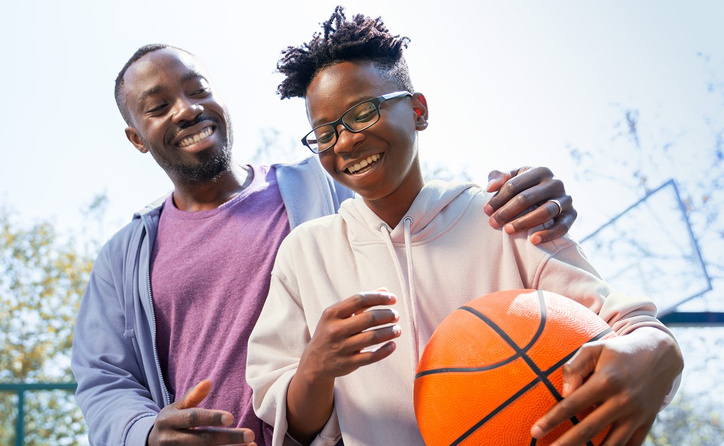 A father and son smiling outdoors and the son is holding a basketball