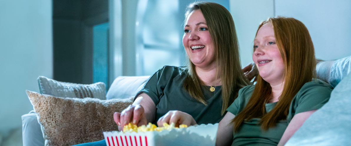 Aubri, a TZIELD® (teplizumab-mzwv) patient, smiling and sitting on the couch eating popcorn with her mother and caregiver, Molly