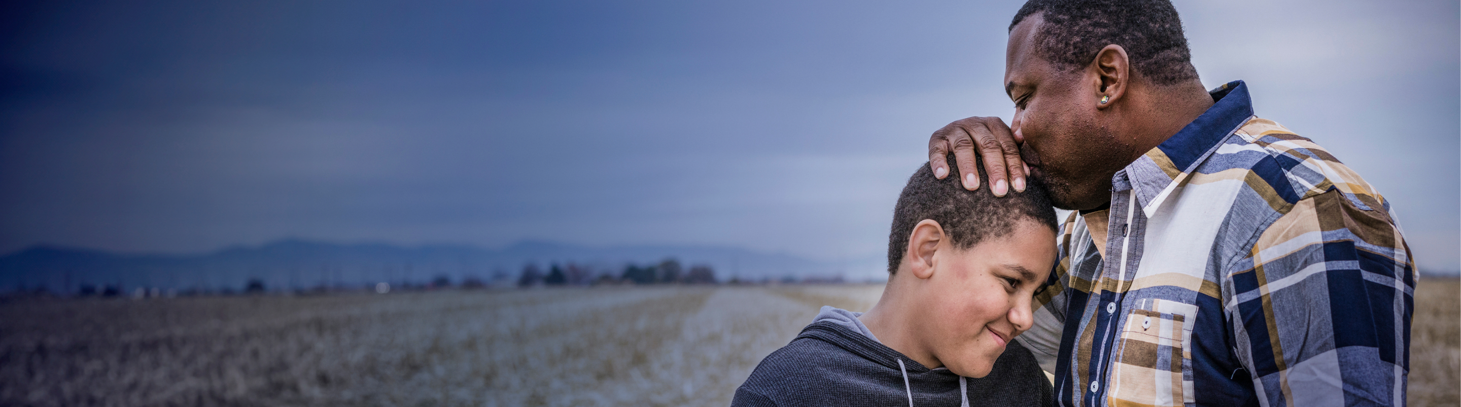 A father holding the top of his son's head and kissing it while standing outdoors