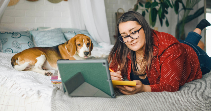 A dog and a woman lounging on a bed while the woman's tablet is propped up and she is writing notes