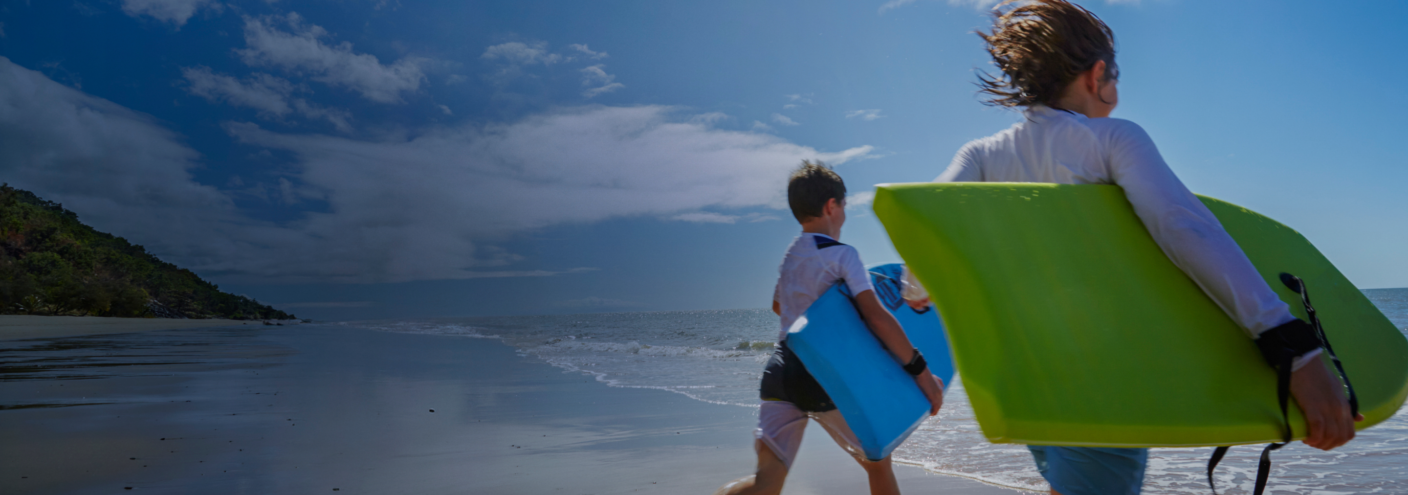 Two young children on a beach running towards the water with bodyboards